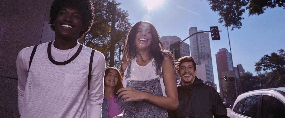 Foto mostra grupo de 4 pessoas andando na rua, sorrindo, em um dia ensolarado