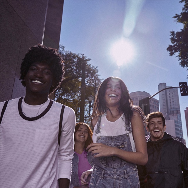 Foto mostra grupo de 4 pessoas andando na rua, sorrindo, em um dia ensolarado