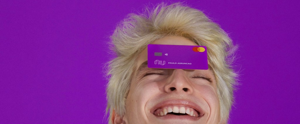 1,5 bilhão de transações header: Foto mostra um jovem em um fundo roxo com um cartão nubank equilibrado na testa