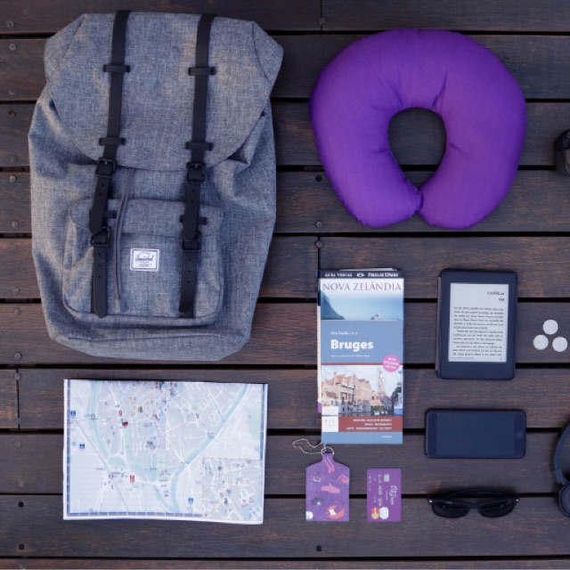 Como planejar uma viagem: vista de cima de uma mochila cinza, um mapa de cidade, um guia de Bruges, um  cartão Nubank e um travesseiro de viagem roxo