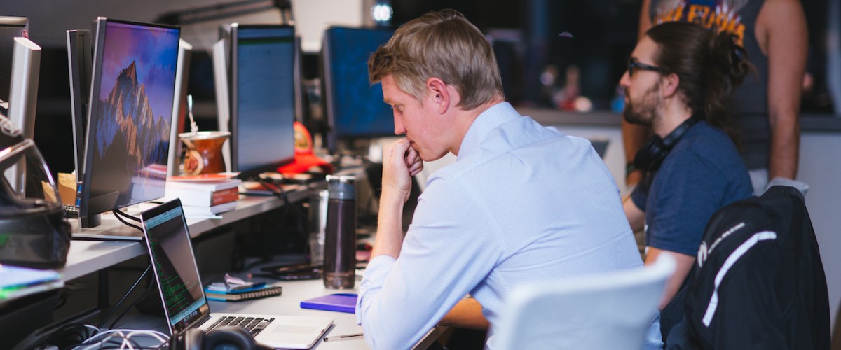 Programação Funcional: no Nubank, um homem está sentado em frente à mesa com o computador aberto. Ele está programando. Atrás, outro homem está sentado e um em pé ao lado dele.