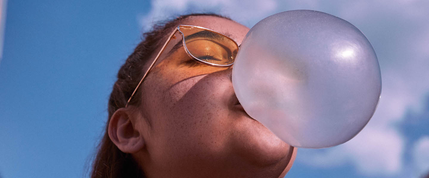 Dia do Solteiro: foto de uma mulher com camiseta preta e óculos de sol amarelo fazendo uma bola de chiclete com a boca.