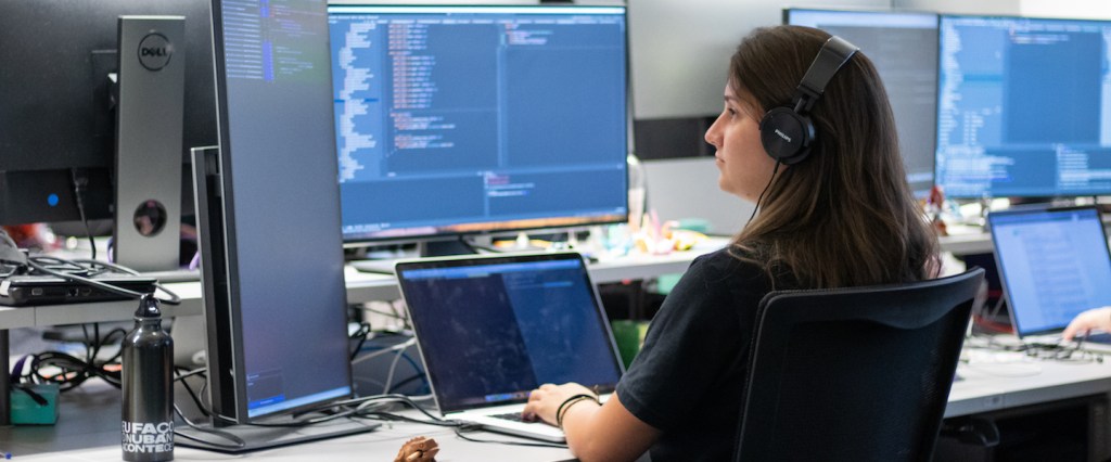 Engenharia de software Nubank: uma mulher com fones de ouvido programando. Ela está com um notebook na mesa e usa dois monitores auxiliares.