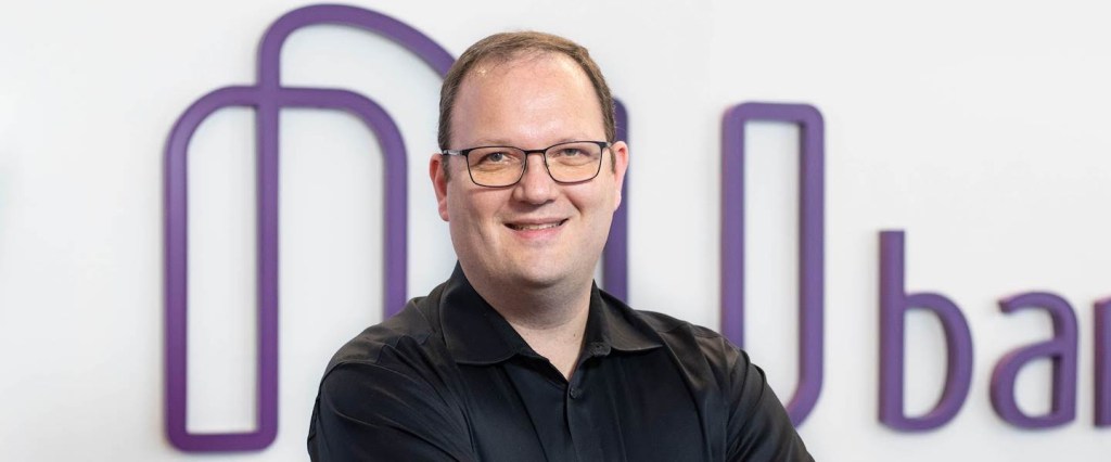 Design de Produto: fotografia de Lucas Pettinati, VP de Design do Nubank, posando em frente ao logo do Nu. Ele veste camisa preta e usa óculos de grau.