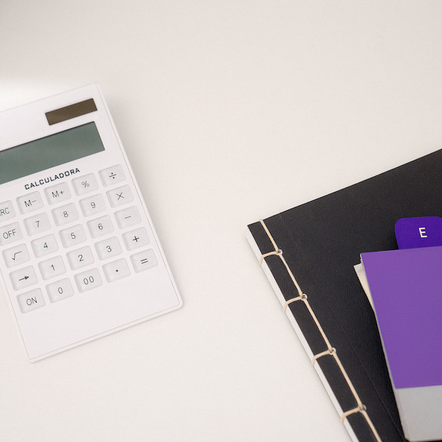Imagem de uma calculadora branca em cima de uma superfície clara. Ao lado, uma pasta preta e umas fichas em tons de lilás e roxo, com documentos.