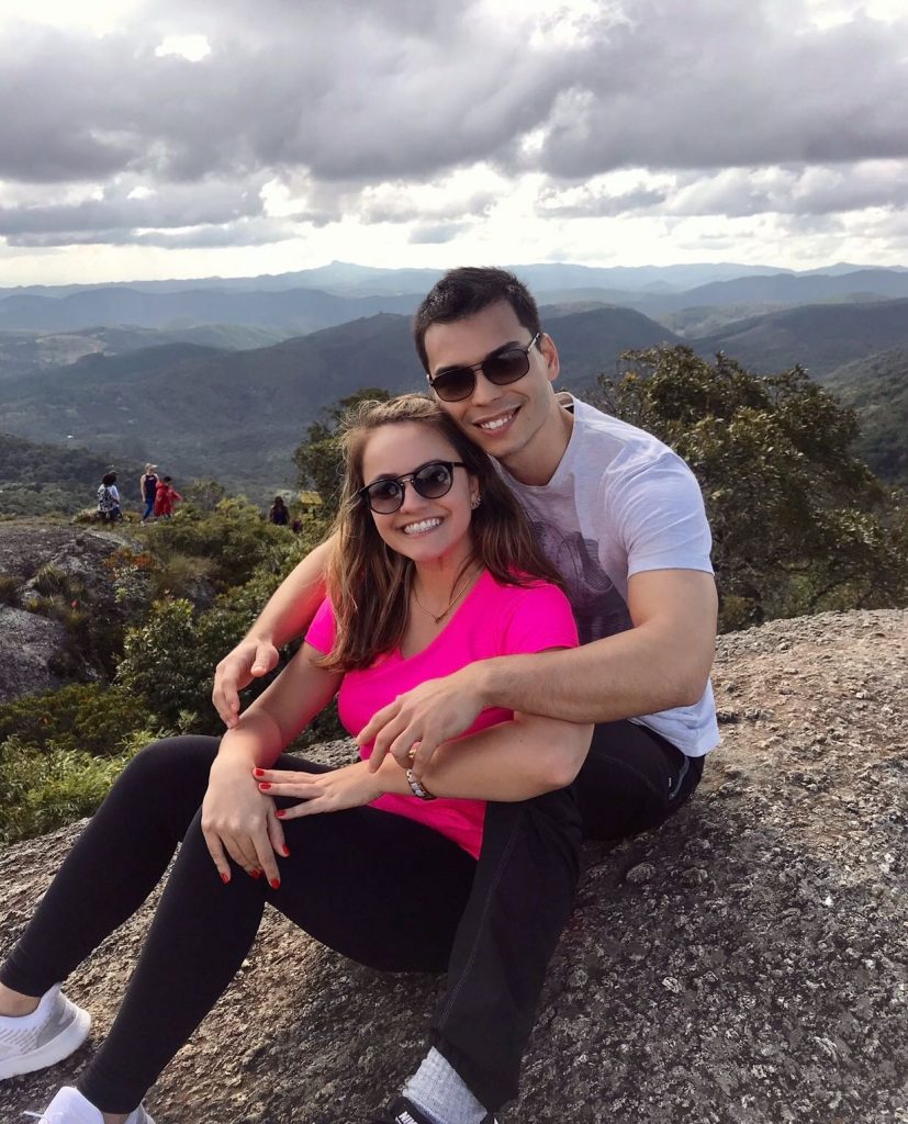 Morar Junto: fotografia de Rafael e Luana sentados abraçados em uma pedra, no alto da montanha, com uma bela vista atrás.