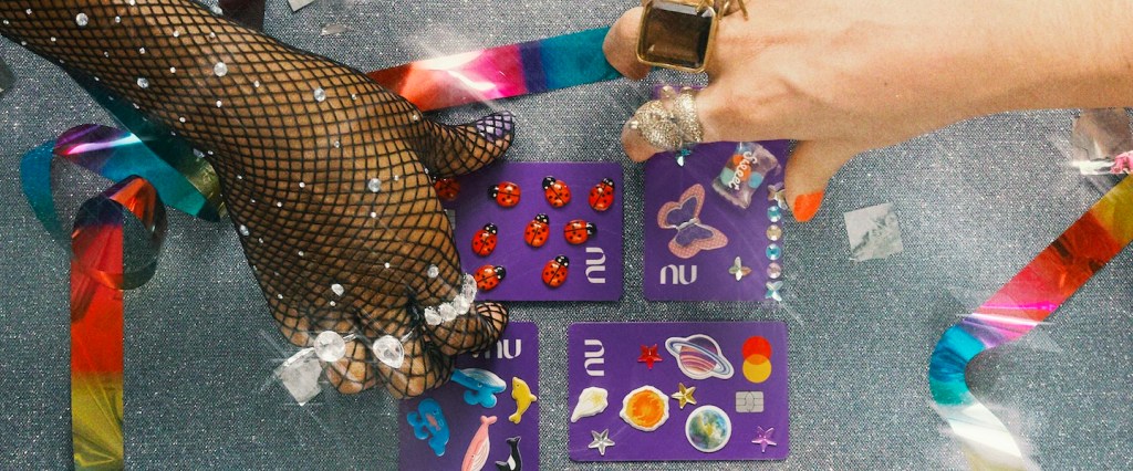 Adesivo para cartão de crédito: imagem de cinco cartões do Nubank adesivados sobre uma superfície prateada, com fitas coloridas em volta. Há também três mãos segurando os cartões, todas elas com anéis e brilhos carnavalescos.
