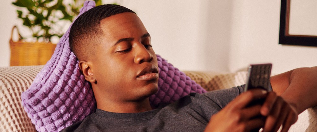 Um homem deitado em um divã com uma almofada lilás e mexendo no celular
