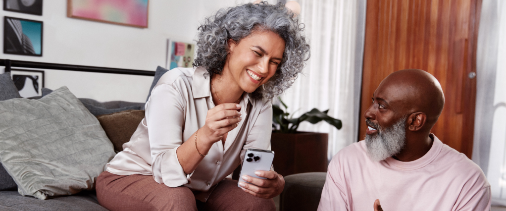 Imagem de uma mulher negra ao lado de um homem negro, e ambos estão sorrindo. Ela segura um celular com uma das mãos e veste uma camisa de seda off white e uma calça de linho marrom. Ela tem cabelos grisalhos, cacheados, na altura do ombro. O homem veste uma camiseta rosa claro, é careca e tem barba branca e comprida. Eles estão em uma sala de estar, com sofá, almofadas, quadros, plantas e uma mesa de centro com objetos decorativos e um prato com frutinhas.