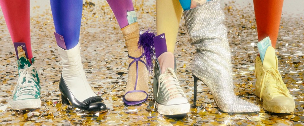 Carnaval: imagem de vários pés vestindo meias calças coloridas nas cores vermelho, azul, amarelo e roxo. Cada pé está com um calçado diferente, sendo três tênis, uma bota de brilho prateada, uma sandália de tiras roxas e um sapatinho preto de lacinho. O chão está forrado de glitter.
