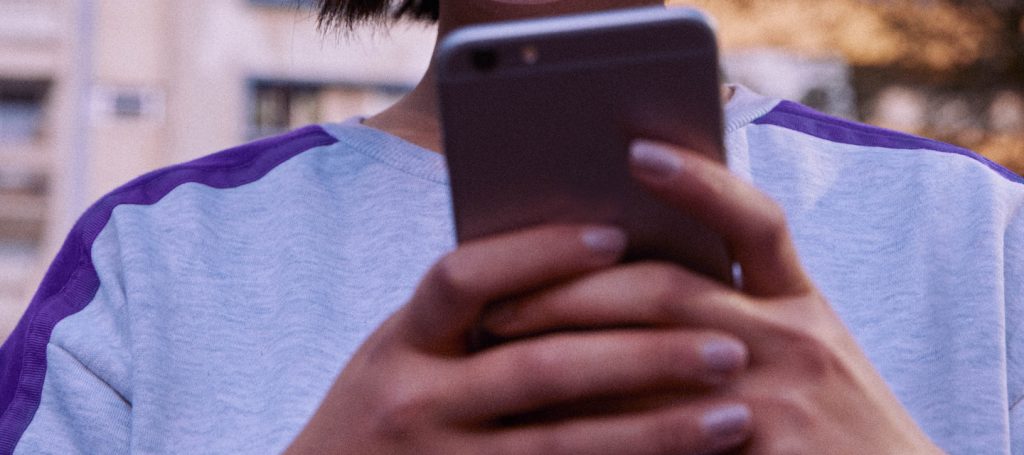 Higienizar celular: garota vestindo moletom cinza e roxo segura um celular na altura do peito.
