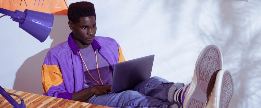 Online: Homem vestindo jaqueta roxa sentado com os pés sobre a mesa e trabalhando em um notebook