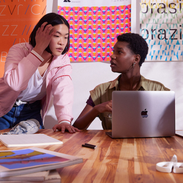 Internet banking: foto mostra duas jovens conversando em frente a uma parede de post its. Uma delas está sentada em uma mesa, com um computador aberto
