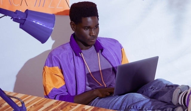 Homem vestindo jaqueta roxa sentado com os pés sobre a mesa e trabalhando em um notebook
