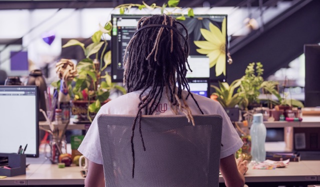 Atendimento Nubank: Jovem com dreads no cabelo sentado, de costas, em uma mesa de trabalho.