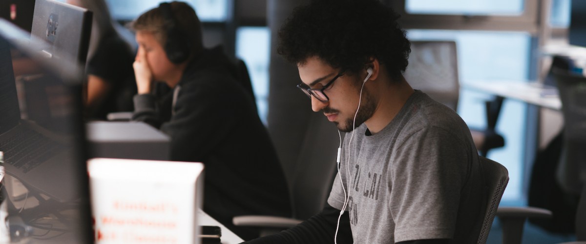 imagem de um homem em primeiro plano, um homem ao seu lado e uma mulher ao fundo, todos trabalhando em seus computadores