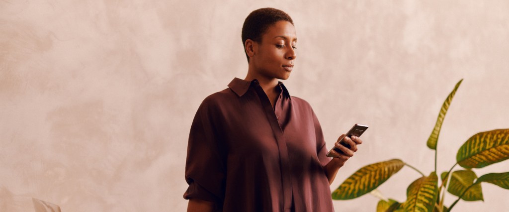 Foto de uma mulher negra, de cabelo bem curto e vestindo uma camisa longa marrom escura. Ela está olhando o celular com uma mão e a outra se apoia em uma mesa onde há um computador e materiais de trabalho. Do outro lado está uma planta, tudo em frente a uma parede neutra
