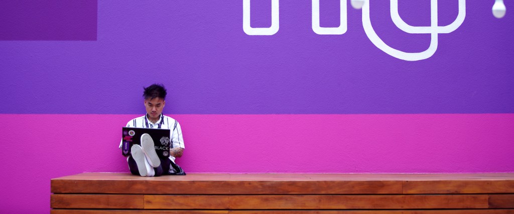 Garoto sentado em um tablado de madeira no canto esquerdo inferior da foto, segurando um notebook. Ele estáem frente aum mural rosae roxo onde está escrito NU em branco.