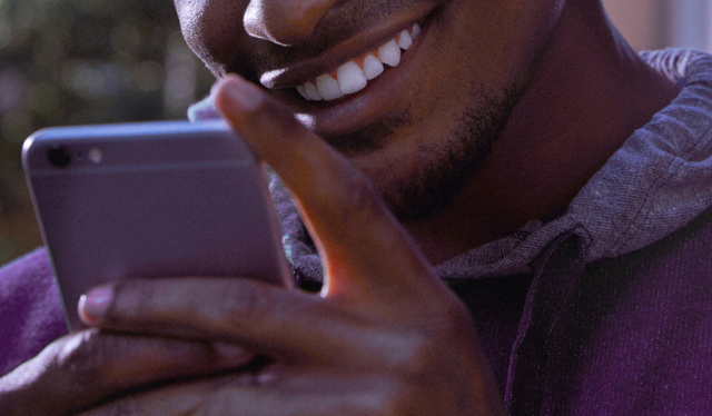Jovem vestindo moletom roxo segura um celular em frente ao seu rosto enquanto sorri.