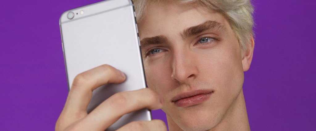 App Nubank vertical: em foto de estúdio com fundo roxo, um homem olha para a tela do celular que ele segura em frente ao rosto.