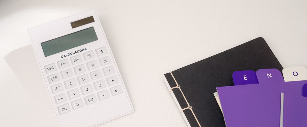 Imagem de uma calculadora branca em cima de uma superfície clara. Ao lado, uma pasta preta e umas fichas em tons de lilás e roxo, com documentos.