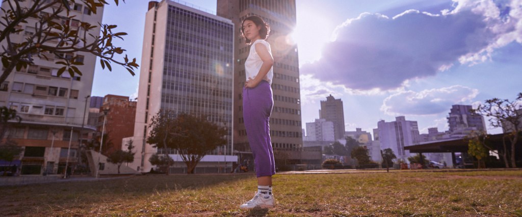 Nubank zera emissão de carbono: fotografia de uma mulher em pé sobre a grama com prédios e um céu azul ao fundo