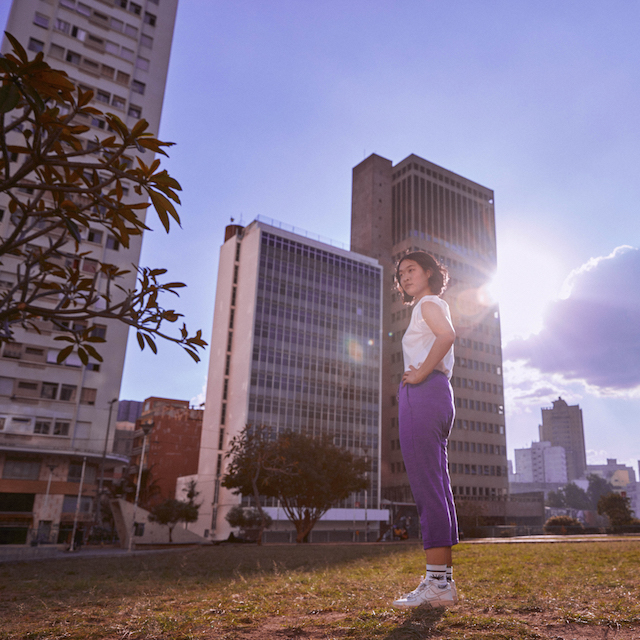 Nubank zera emissão de carbono: fotografia de uma mulher em pé sobre a grama com prédios e um céu azul ao fundo