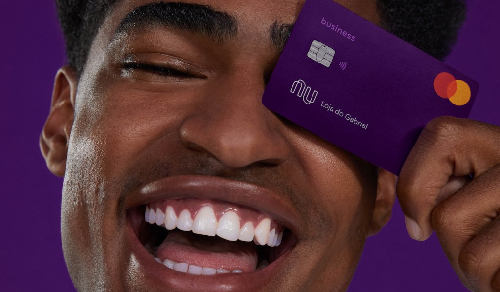 Como funciona o débito na conta PJ Nubank: foto em estúdio com fundo roxo de um homem sorrindo segurando o cartão PJ em frente ao olho esquerdo