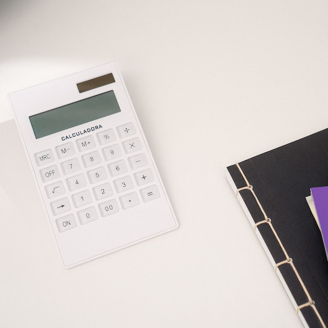 Imagem de uma calculadora branca ao lado de um caderno preto e de etiquetas roxas sobre uma mesa de escritório branca.