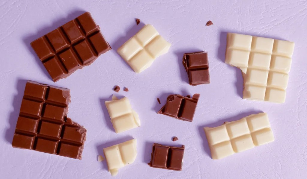 Drawback: Foto de vários quadradinhos de chocolate branco e ao leite espalhados por um fundo lilás