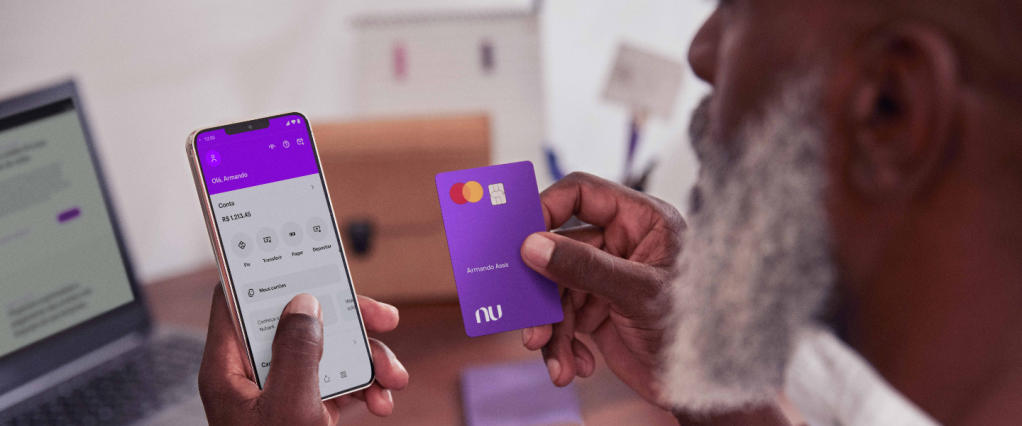 Homem negro de barba branca segura o cartão roxinho do Nubank e uma mão e um celular, com a página inicial da conta do Nubank na outra mão.