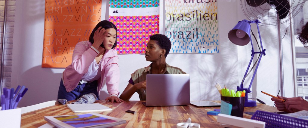 Empreendedorismo feminino no Brasil: foto mostra duas jovens conversando em frente a uma parede de post its. Uma delas está sentada em uma mesa, com um computador aberto