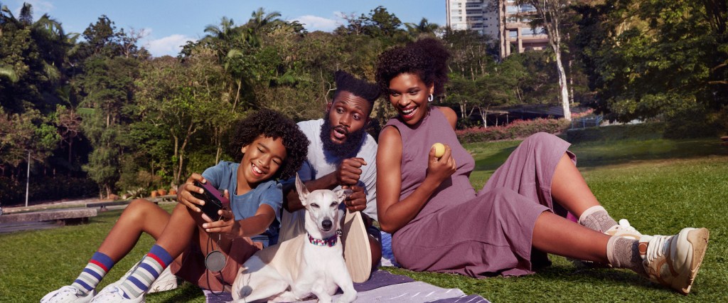Imagem de uma família negra, com um cachorro. Eles estão em um parque, sentados numa toalha em tons de roxo.