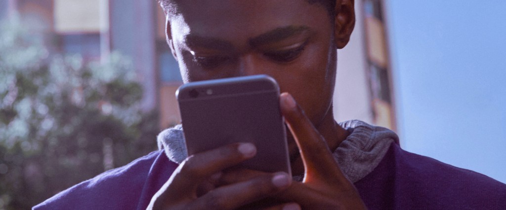 App Nubak 5o colocado: jovem vestindo moletom roxo segura um celular em frente ao seu rosto enquanto sorri.
