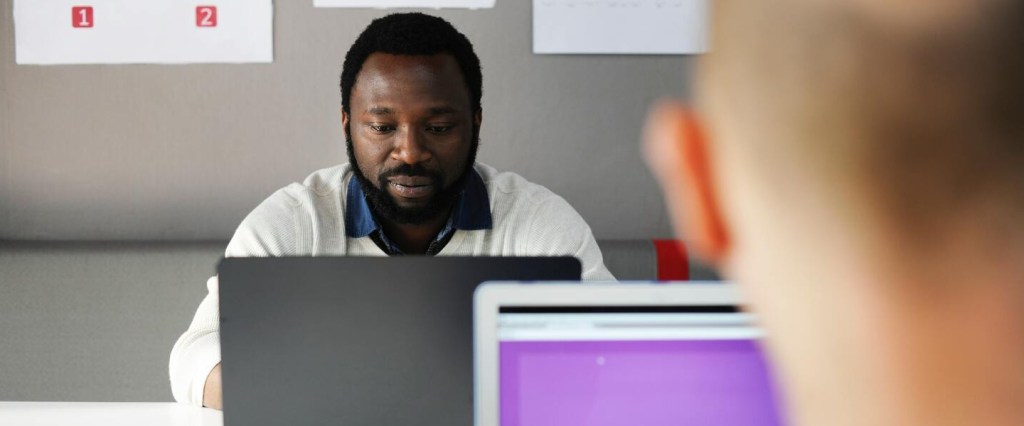 homem negro trabalhando com um notebook em frente a um homem branco que está de costas também com um notebook exercendo suas profissões