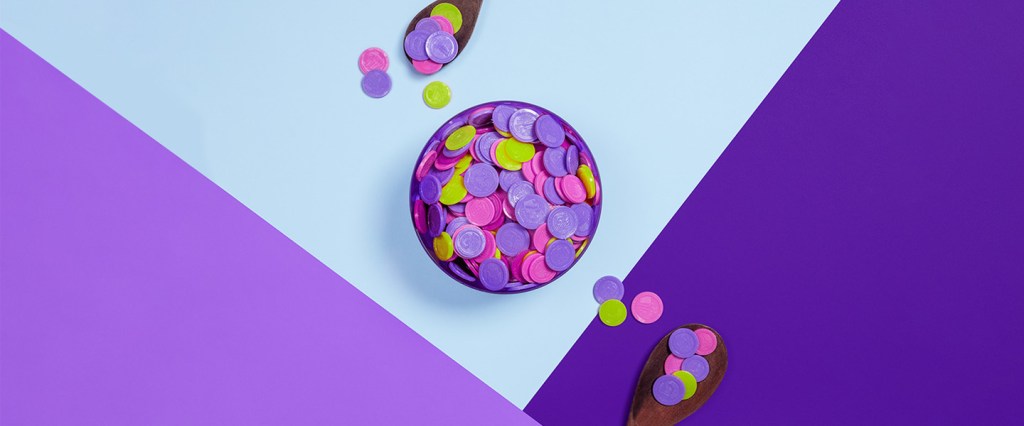 Rendimento da Poupança - Imagem de um pote cheio de moedas coloridas, com duas colheres ao lado dele. O pote está sobre uma mesa em tons de roxo e azul.