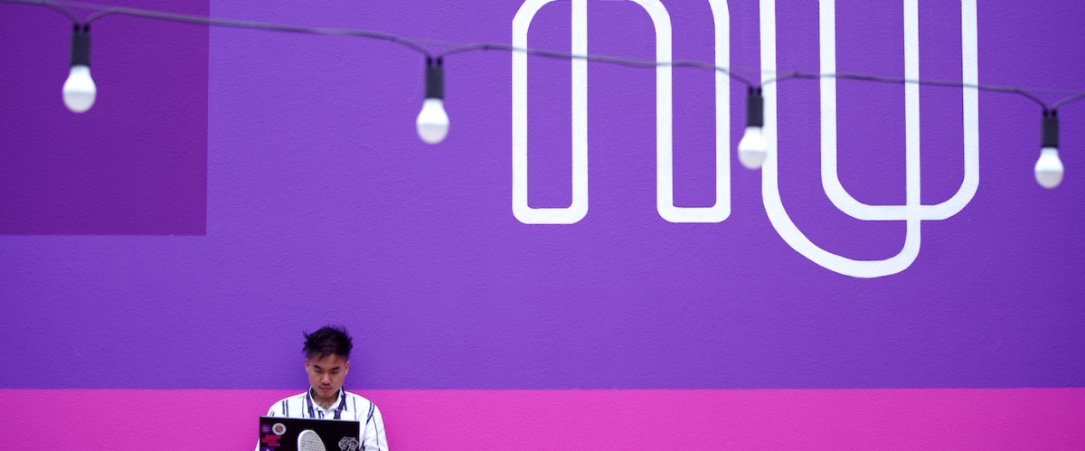 Garoto sentado em um tablado de madeira no canto esquerdo inferior da foto, segurando um notebook. Ele está em frente a um muro rosa e roxo onde está escrito NU em branco.