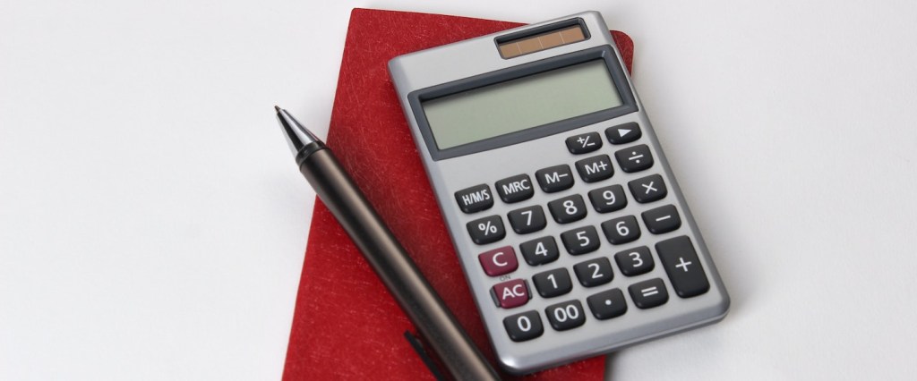 Tabela imposto de renda 2021: sobre uma mesa branca, repousam uma calculadora cinza e uma caneta preta sobre um caderninho vermelho