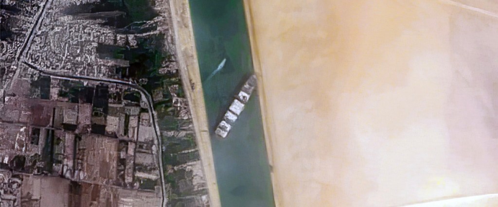 Foto aérea mostra navio na diagonal bloqueando a passagem do canal de Suez. ã esquerda, casas. À direita, areia.