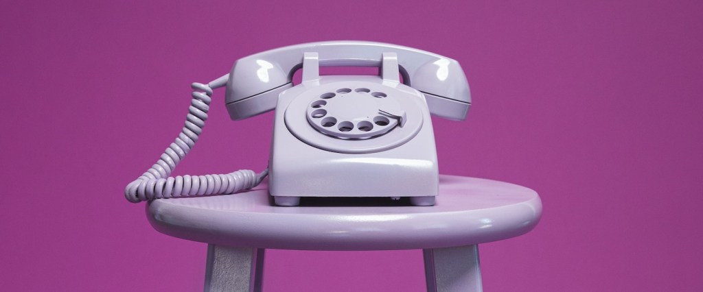 Pix lista de contatos celular: fotografia de um telefone analógico branco, sobre um banco roxo num fundo roxo