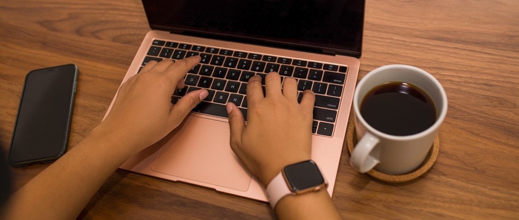imagem de mãos de mulher digitando em um notebook com uma xícara de café e um celular ao lado