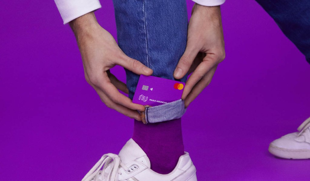 Foto mostra um cartão Nubank colocado dentro de uma meia roxa por um jovem usando um tênis branco.