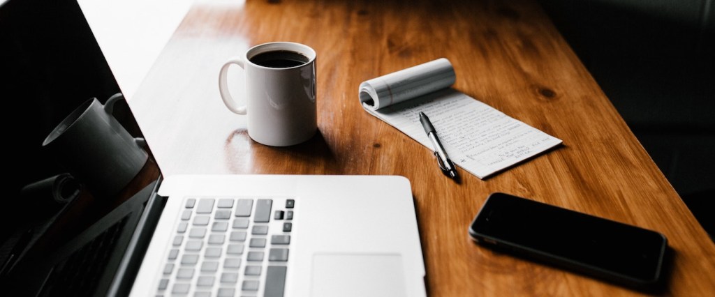 Como se cadastrar no INSS: fotografia de um notebook, uma xícara branca com café, um celular, um bloco de papel e uma caneta apoiados sobre uma mesa de madeira em frente a uma janela