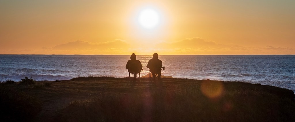 INSS no Imposto de Renda 2021: fotografia de duas pessoas sentadas num morro, à beira do mar, assistindo o sol se pôr.