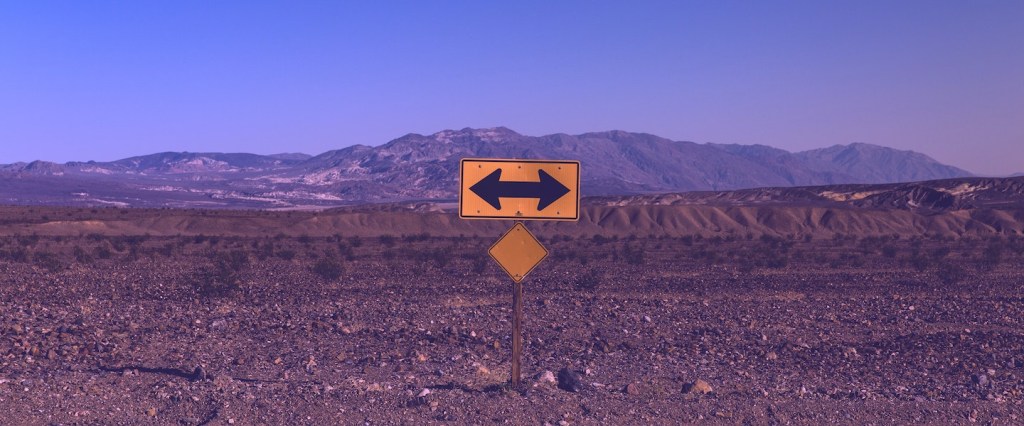 O que é pivotar significado: fotografia de uma placa amarela com uma seta apontado para a esquerda e para a direita. Ao fundo, uma montanha e um terreno árido.