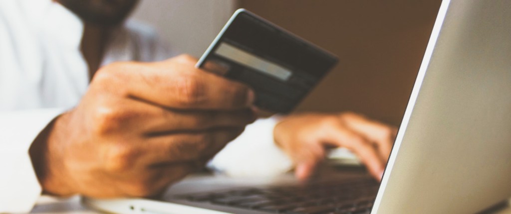 Semana do Consumidor: fotografia de um homem segurando um cartão de crédito com a mão direita enquanto digita num notebook com a mão esquerda