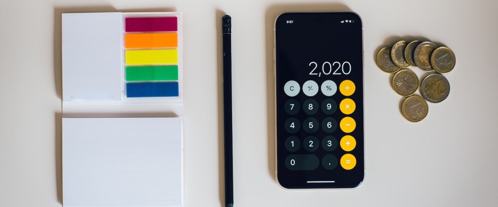 Fotografia de itens dispostos sobre uma mesa branca: um celular com a calculadora aberta, uma pilha de moedas, um lápis e um caderninho de anotações com post its coloridos