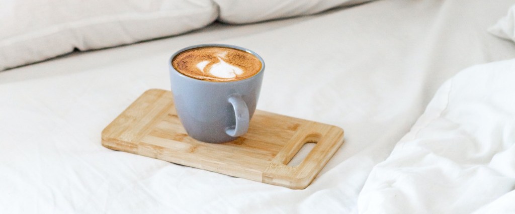 Pix funciona final de semana: fotografia de uma xícara de café cinza, com café dentro, sobre um suporte de madeira em cima de uma cama com lençol e travesseiros brancos