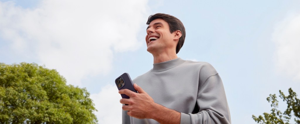 Como investir no Nubank: imagem de um homem branco segurando um celular em um ambiente externo, com árvores e céu azul. Ele veste uma blusa cinza de manga comprida e está sorrindo.