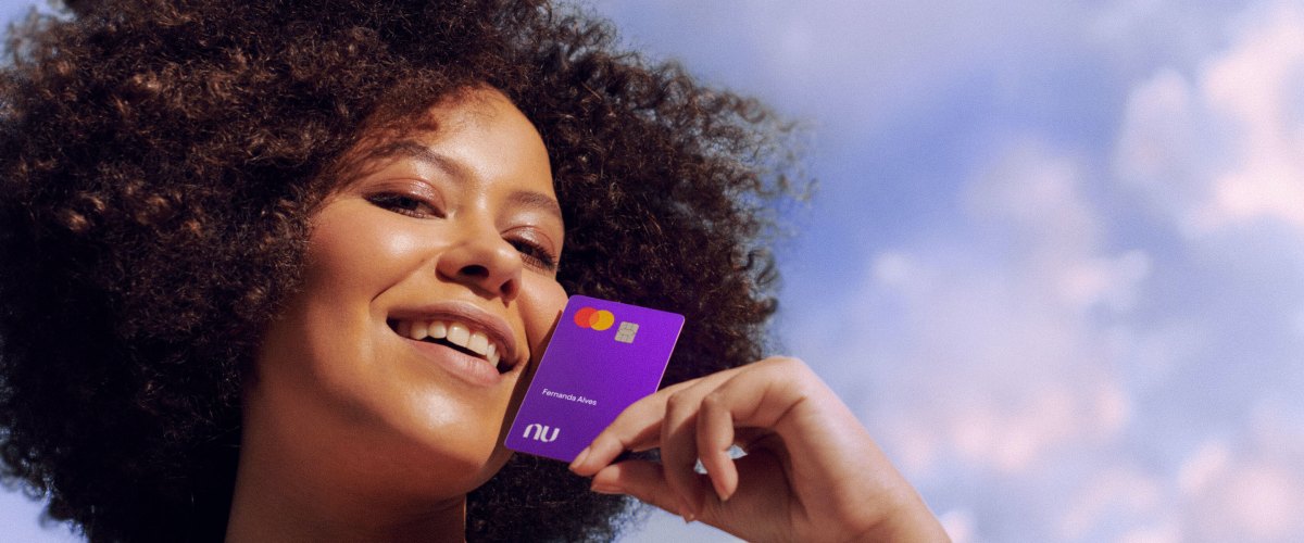 imagem de uma mulher sorrindo, olhando para a câmera de cima para baixo. Ela segura um cartão roxo do Nubank